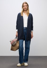 Granatowy kardigan z otwartym przodem, biała koszula zapinana na guziki, niebieskie dżinsy oraz brązowa torba z wzorzystym szalikiem zarzuconym na nią.