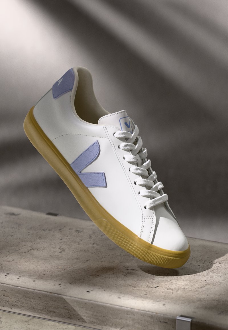 Balti sneakers ar lavandas akcentiem, apaļš purngalis, plakani auklas un teksturēta gumijas zole. Sānu daļā ir liels "V" logotips no zamšādas.