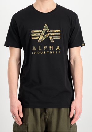 Homme portant un t-shirt noir avec un logo Alpha Industries camouflage et du texte, associé à un pantalon cargo vert olive.