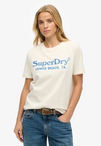 Kremowa bawełniana koszulka z niebieskim napisem "SuperDry" i "VENICE BEACH. CA." Krótkie rękawy, luźny krój, noszona z niebieskimi dżinsami i brązowym paskiem.