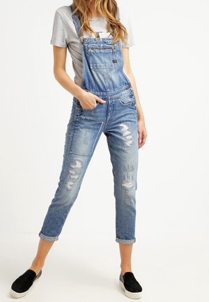 Jasnoniebieskie ogrodniczki z przetarciami. Posiadają regulowane ramiączka, detale z guzikami oraz kwadratową kieszeń. Noszone na szarym t-shircie.