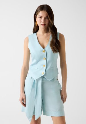 Femme portant un gilet sans manches bleu clair avec des boutons dorés et un short taille haute assorti avec une longue ceinture en tissu, debout devant un fond blanc.