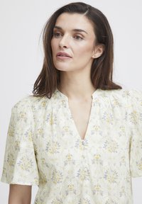 Blommig blus i ljusgul med V-ringning, korta puffärmar och intrikata mönster av blå och gula blommor. Mjuk, texturerad tyg.