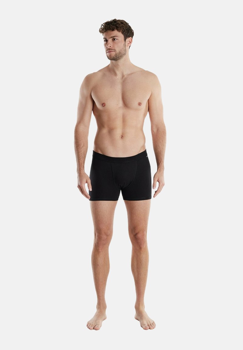 Svarta boxershorts gjorda av elastiskt tyg, med en åtsittande design och elastisk midjeband. Modellen står barfota och visar en neutral hållning.