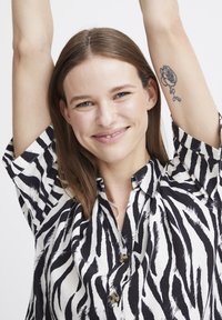 Camicia a righe zebrate in bianco e nero con maniche corte, chiusura a bottoni e colletto. Presenta un tessuto liscio e una leggera lucentezza.