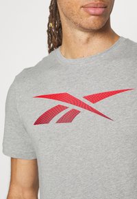 Detailní záběr osoby v šedém tričku s velkým červeným logem Reebok na hrudi a viditelnými dreadlocky.