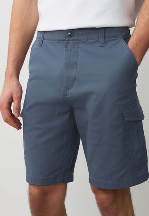 Mand iført blå cargoshorts med sidelommer og hænder hvilende tæt på lommerne, kombineret med en hvid skjorte.