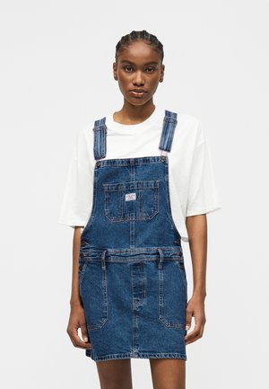 Kobieta w białej koszulce pod niebieską dżinsową sukienką z kieszeniami z przodu, stojąca na jednolitym jasnym tle.