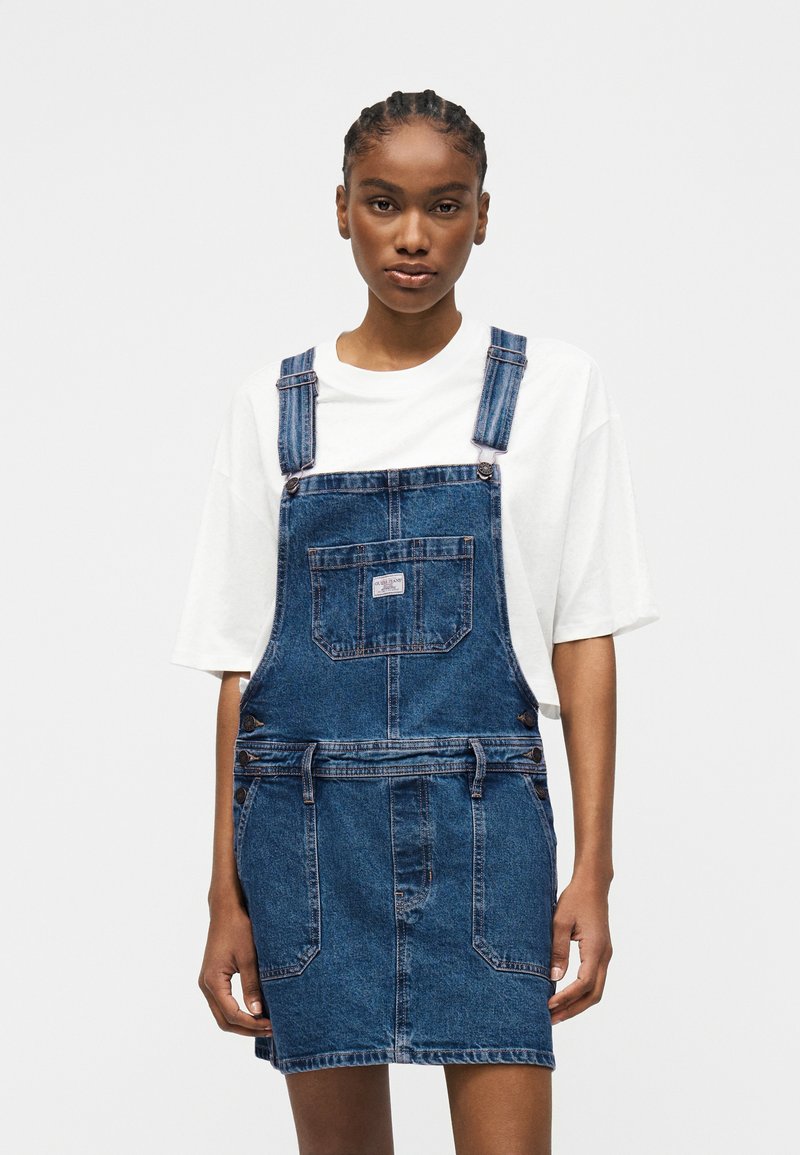 Kobieta w białej koszulce pod niebieską dżinsową sukienką z kieszeniami z przodu, stojąca na jednolitym jasnym tle.