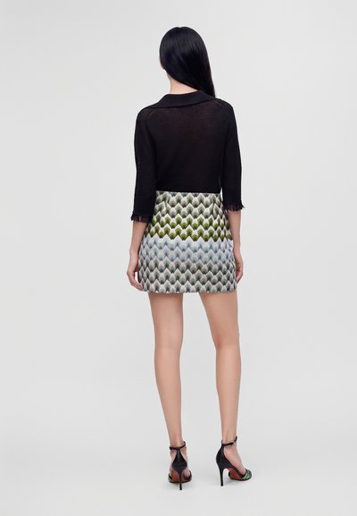 Femme aux longs cheveux noirs, portant un haut noir et une mini-jupe à motifs, debout de dos sur un fond gris clair uni.