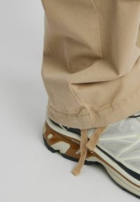 Pantalon beige avec un bas retroussé, associé à une sneaker gris clair avec un dessus texturé, des accents noirs et un détail réfléchissant.