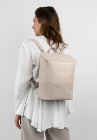 Beige läderryggsäck med en slät textur, dragkedjestängning och en kontrasterande mockabotten. Minimalistisk design med justerbara remmar.