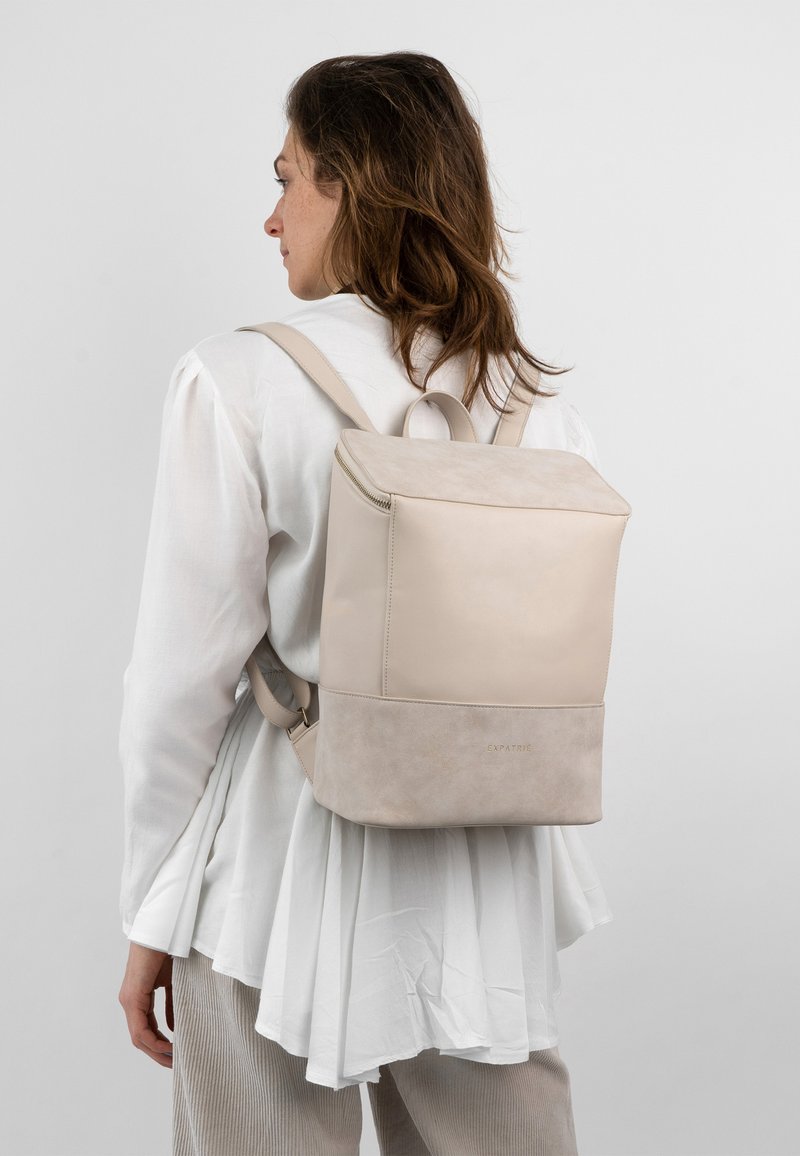 Beige läderryggsäck med en slät textur, dragkedjestängning och en kontrasterande mockabotten. Minimalistisk design med justerbara remmar.