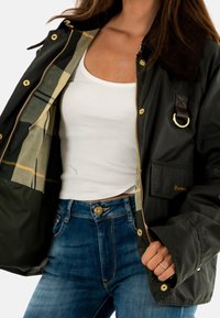 Dunkelgrüne gewachste Jacke mit einem Cordkragen, goldenen Beschlägen, kariertem Futter und einer Fronttasche, die dezente Markenkennzeichnung in Gold aufweist.