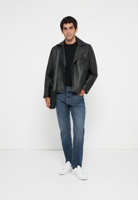 Joven con chaqueta de cuero negra, camiseta negra, vaqueros azules, zapatillas blancas y llevando un bolso de hombro negro, de pie contra un fondo blanco.