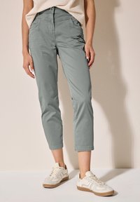 Pantaloni cropped verdi con una texture liscia, dotati di tasche frontali e dettagli sulle cuciture. Abbinati a sneakers di colore chiaro.
