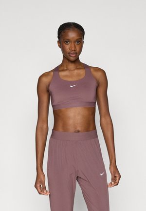 Brassière de sport Nike et pantalon taille haute assorti dans une teinte mauve douce. La brassière présente des bretelles épaisses, un accent de logo et des détails subtils de texture.