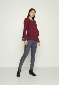 Blusa de maternidade em burgundy, de mangas longas com franzidos laterais. Combinada com jeans skinny cinza escuro e botas de tornozelo pretas. Inclui uma bolsa cruzada preta.