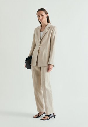 Mujer con traje beige a medida, con bolso negro de mano, lleva una camiseta blanca y sandalias negras de tacón, de pie frente a un fondo claro liso.