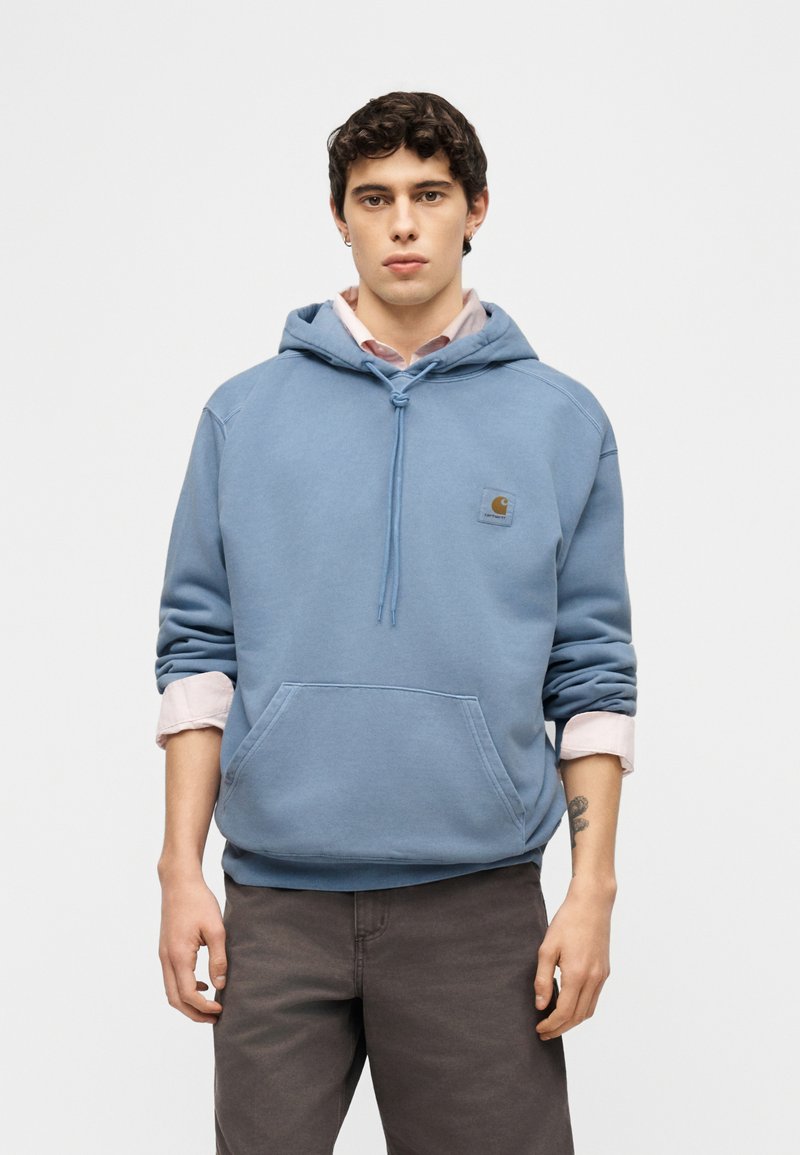 Junger Mann mit lockigem Haar, der einen blauen Carhartt-Hoodie über einem hellrosa Hemd trägt und vor einem schlichten hellen Hintergrund steht.
