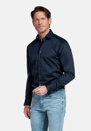 Man in een nauwsluitend donkerblauw overhemd met knopen en lichtblauwe spijkerbroek, staand tegen een effen witte achtergrond.