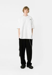 Camiseta oversized blanca con detalles de corte únicos, combinada con pantalones negros de corte suelto que cuentan con un acento de cadena y zapatos texturizados.