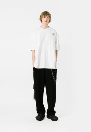 Camiseta oversized blanca con detalles de corte únicos, combinada con pantalones negros de corte suelto que cuentan con un acento de cadena y zapatos texturizados.