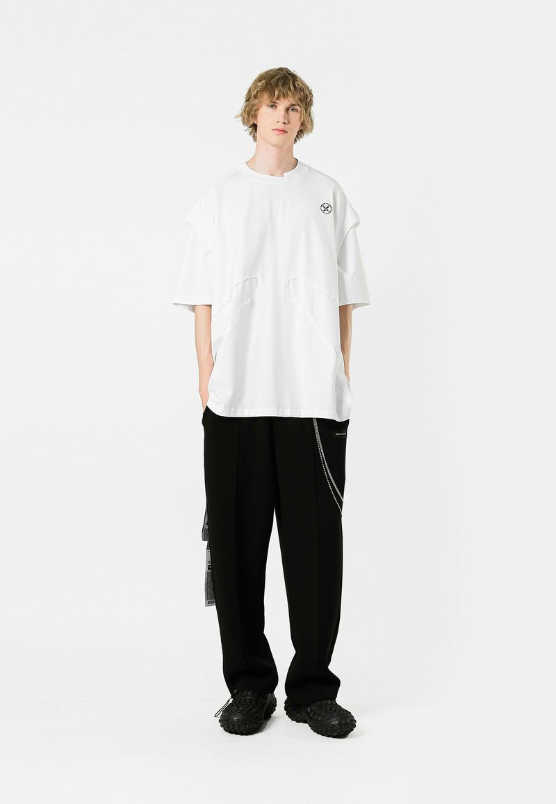 Camiseta oversized blanca con detalles de corte únicos, combinada con pantalones negros de corte suelto que cuentan con un acento de cadena y zapatos texturizados.
