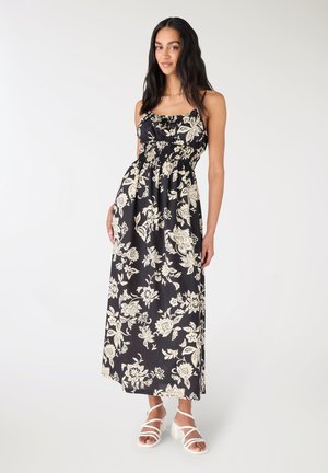 Donna che indossa un vestito lungo senza maniche nero con motivi floreali bianchi e sandali bianchi con tacco a blocco e cinturini, in piedi su uno sfondo bianco.