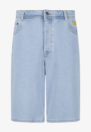 Homme portant un short en jean bleu clair jusqu’aux genoux, des chaussettes blanches et des baskets blanches, debout, les mains dans les poches avant devant un fond uni.