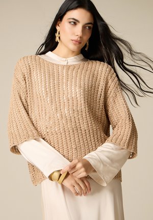 Kvinde med langt mørkt hår iført beige hæklet top over en cremefarvet bluse, guldfarvede søstjerne-øreringe og et guldbånd mod en neutral baggrund.