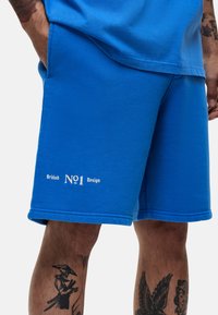 Shorts en coton bleus avec une coupe décontractée, dotés de poches latérales et d'un texte brodé "British No1 Design" sur l'ourlet inférieur.