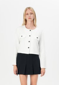 Weiße, gekürzte Jacke aus strukturiertem Stoff mit zwei vorderen Taschen, schwarzen Knöpfen und langen Ä sleeves, kombiniert mit einem schwarzen, plissierten Rock.