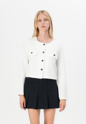 Hvit croppet jakke med en teksturert stoff, med to frontlommer, svarte knapper og lange ermer, kombinert med et svart plisserte skjørt.
