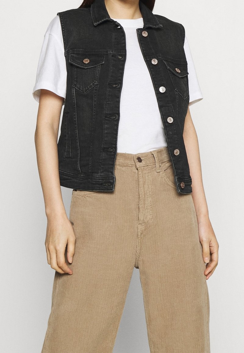 Person klädd i svart utanärmad denimjacka över vit kortärmad skjorta och beige corduroybyxor, stående mot en enkel bakgrund.