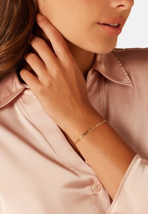 Gouden schakelarmband om een pols, met kruisende schakels. De hand steunt tegen een lichtroze satijnen shirt met een kraag en knoopdetail.