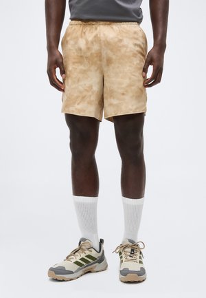 Mann trägt beige Batik-Shorts, weiße Knöchelsocken und beige-grau-grüne Sneakers, steht vor schlichtem Hintergrund.