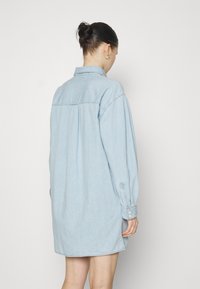 Abito camicia in denim azzurro chiaro con maniche lunghe, polsini a bottoni, colletto e vestibilità ampia, caratterizzato da una piega sul retro per una maggiore libertà di movimento.