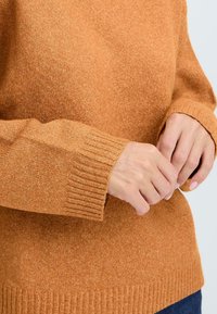 Pullover in un caldo arancione bruciato, realizzato con un finissaggio testurizzato e con orli e polsini a costine. Il tessuto sembra morbido e leggermente macchiato.