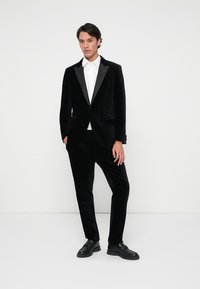 Zwarte fluwelen blazer met satijnen revers, gecombineerd met op maat gemaakte broeken. Witte blouse eronder, model draagt zwarte schoenen. Effen witte achtergrond.