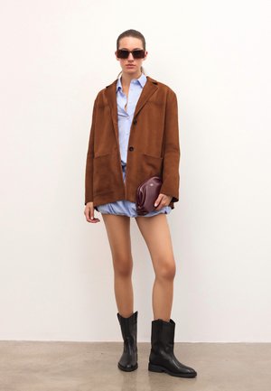 Kobieta w oversize'owej brązowej kurtce, jasnoniebieskiej mini sukience, czarnych botkach do połowy łydki, w ciemnych okularach przeciwsłonecznych, trzymająca małą bordową torebkę.