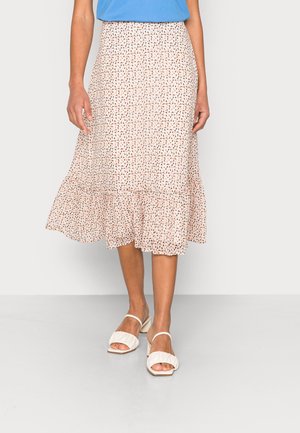 Femme portant une jupe mi-longue rose clair à imprimé floral avec un ourlet à volants et des sandales blanches à bouts ouverts, debout devant un fond uni.
