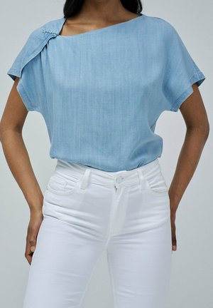 Femme portant un chemisier ample bleu clair à manches courtes avec un pli sur l'épaule et un jean blanc taille haute ajusté.