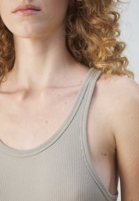 Gros plan d'une personne aux cheveux châtain clair bouclés, portant un débardeur côtelé beige, montrant le cou, la clavicule et la partie supérieure de la poitrine.
