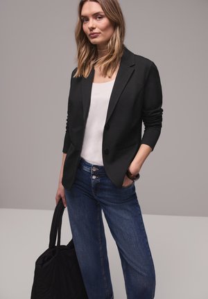 Femme portant un blazer noir, un haut blanc, un jean bleu, tenant un sac cabas noir, debout devant un fond gris uni.