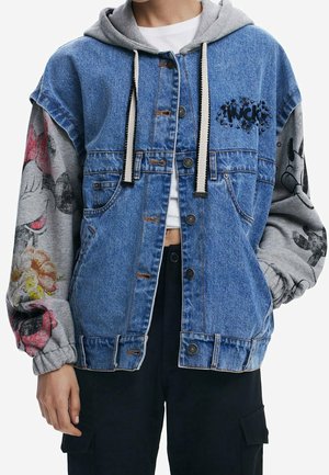 Personne portant un gilet en jean bleu avec un sweat à capuche gris en dessous, orné de graphismes de dessins animés sur les manches, et un pantalon noir.