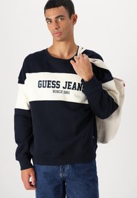 Navy sweatshirt met een witte horizontale strook, "GUESS JEANS" logo, lange mouwen en geribbelde mouwen. Gecombineerd met denim jeans en een beige tas.