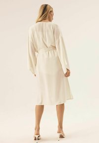 Długa, kremowa sukienka z długimi rękawami, talią podkreśloną paskiem i swobodnym krojem. Gładki materiał o lekkiej strukturze.