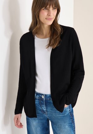 Nainen pukeutuneena mustaan avoimeen neuletakkiin, valkoisen paidan ja sinisten farkkujen kanssa, seisoo yhdellä kädellä taskussa neutraalia taustaa vasten.