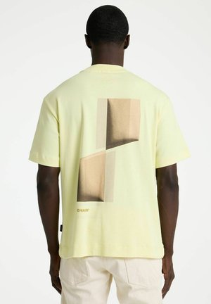 Man draagt een lichtgeel T-shirt met korte mouwen met een abstracte beige en bruine geometrische print op de achterkant, gecombineerd met lichte broek.